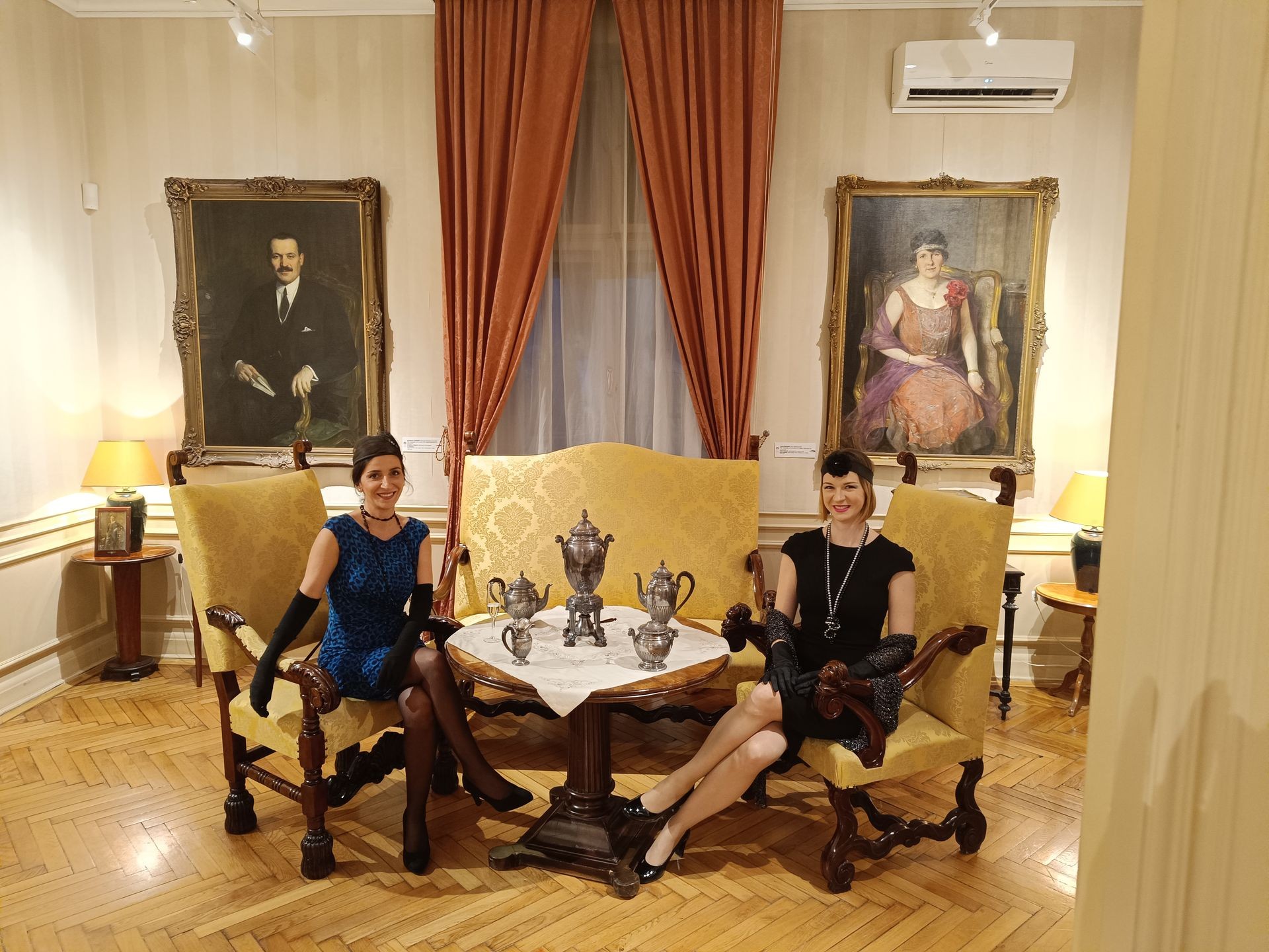 Two women dressed in vintage attire sit in an elegant room with classic portraits and antique furniture.